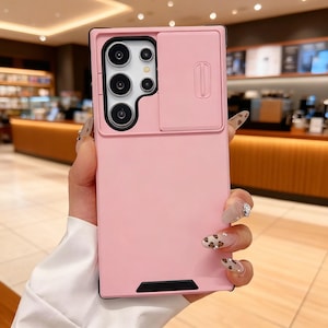 May include: A pink phone case with a sliding camera cover. The case has a rectangular design with a black border and a cutout for the camera lenses. The camera cover is a lighter shade of pink and has a vertical slot. The case is held by a person.