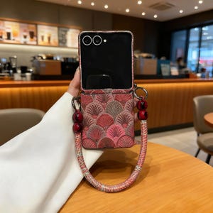 Peut inclure: Un étui de téléphone à motifs rouges et roses avec une sangle assortie. L'étui présente un motif décoratif et une sangle perlée avec des perles rouges. L'écran du téléphone est noir.