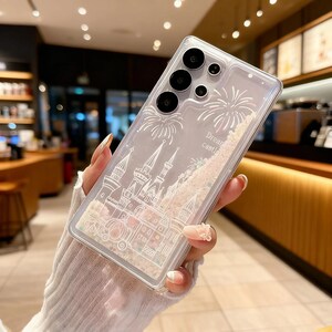 May include: Clear phone case with a castle design and the words "Dream castle". The case has a glittery, liquid-filled section with a castle illustration and fireworks. The phone case is held in a person's hand.