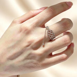 Könnte beinhalten: Ein silberner Ring mit einem einzigartigen Design. Der Ring hat eine käfigartige ovale Form mit einer herabhängenden Kette und einem kleinen, runden Anhänger. Der Ring wird an einem Finger getragen und zeigt seine komplizierten Details und seinen eleganten Stil.