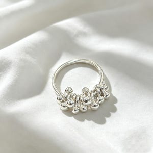 May include: A polished silver ring featuring a series of small, round, metallic beads suspended from the main band. The beads are arranged in a cluster along the lower portion of the circular band, creating a textured, dangling effect in bright silver.