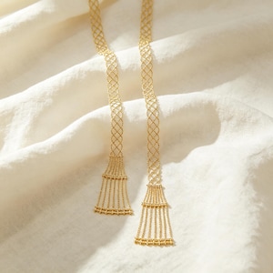 Puede incluir: Un collar de tono dorado con dos cadenas largas y delgadas que presentan un delicado patrón de celosía en forma de diamante. Cada cadena termina en un fleco acampanado en forma de abanico compuesto por múltiples hilos verticales con pequeñas cuentas texturizadas.