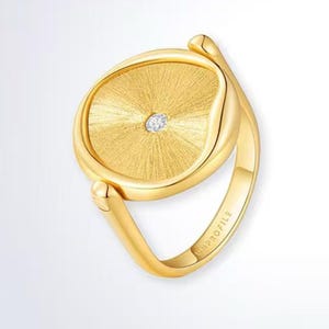 May include: A gold ring featuring a textured, circular face with a central diamond. The ring has a polished band and a distinctive design. The word "UNPROFILE" is engraved on the band.
