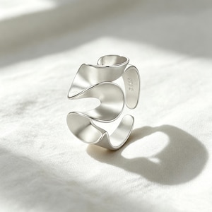 May include: A silver-colored open ring featuring a fluid, wavy, ribbon-like design with smooth, curved edges. The metal surface has a soft, matte finish, and the inner band is clearly stamped with the text 925, indicating its sterling silver composition.