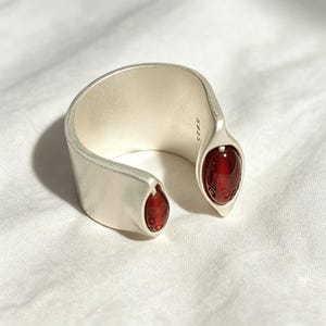 May include: A wide, open-ended silver ring with a matte finish, featuring two marquise-shaped, deep red cabochon stones set at each terminal end. The interior of the band is clearly stamped with the hallmark S925, indicating sterling silver composition.