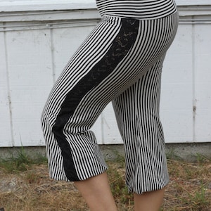 May include: Cropped black and white striped pants with a wide waistband. The pants feature a black lace panel down the side of each leg. The fabric has vertical stripes, creating a slimming effect. The pants are worn at the mid-calf.