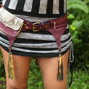 Pu&ograve; includere: Marsupio in pelle bordeaux con patta in pelle di serpente e dettagli dorati. La cintura ha una chiusura con fibbia e una tasca con cerniera. Nappe dorate pendono dal basso. Il marsupio &egrave; indossato sopra un capo a righe nere e argento.