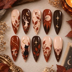 Op de afbeelding: Een set van tien amandelvormige press-on nagels met herfstthema in tinten bruin, crème en roest. De ontwerpen bevatten illustraties van een hert, een zwarte kat, paddenstoelen, dennenbomen, maansikkels, sterren en herfstbladeren.