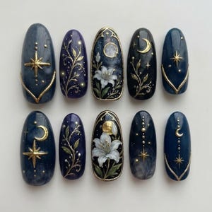 May include: A set of ten oval-shaped press-on nails featuring dark blue and purple backgrounds with intricate gold celestial motifs, including stars and crescent moons, alongside detailed white lily floral illustrations and delicate vine patterns.