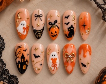 Może przedstawiać: Zestaw dziesięciu sztucznych paznokci w kształcie migdała z motywami halloweenowymi, takimi jak duchy, dynia, czarny kot, nawiedzony dom, nietoperze, pajęczyny i pomarańczowa kratka, ozdobione złotymi zawieszkami, gwiazdkami i małymi perłami.