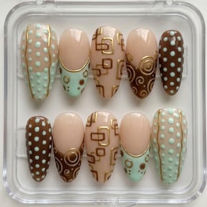 May include: A set of ten artificial nails in a clear plastic case. The nails feature various designs, including polka dots, geometric patterns, and swirls, in shades of brown, beige, and mint green, accented with gold details. The nails are oval-shaped.