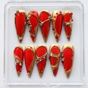 May include: A set of ten red and gold press-on nails. Each nail has a vibrant red base, with gold trim and embellished with rhinestones and small red beads. The nails are displayed in a clear plastic case.