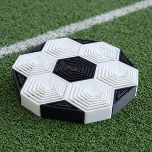 Può includere: Un sottobicchiere stampato in 3D a forma di pallone da calcio. Il sottobicchiere presenta un motivo esagonale bianco e nero, che ricorda il design di un pallone da calcio. È posizionato su una superficie verde con una linea bianca.