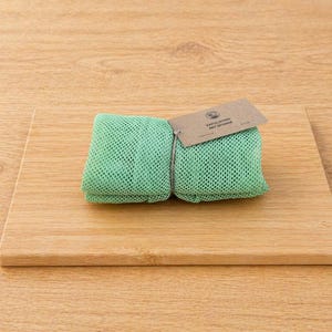 Op de afbeelding: Een paar opgevouwen, lichtgroene exfoliërende netsponges, samengebonden met touw, rustend op een lichtbruine houten snijplank. Een klein, bruin papieren label is aan de sponzen bevestigd.