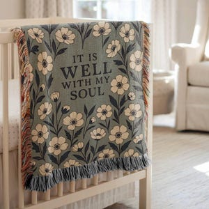 May include: A sage green baby blanket with a floral pattern and the text "IT IS WELL WITH MY SOUL." The blanket is draped over a white cot. The blanket has a fringed edge with red, orange, and tan threads.