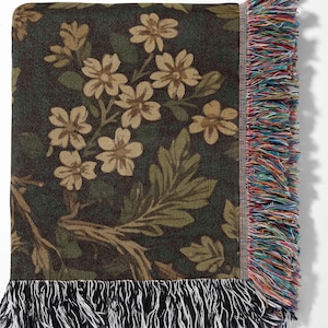 May include: A woven throw blanket featuring a dark green background with a floral pattern of light brown flowers and green leaves. The blanket has a multicolored fringe on the right side and a black and white fringe along the bottom edge.