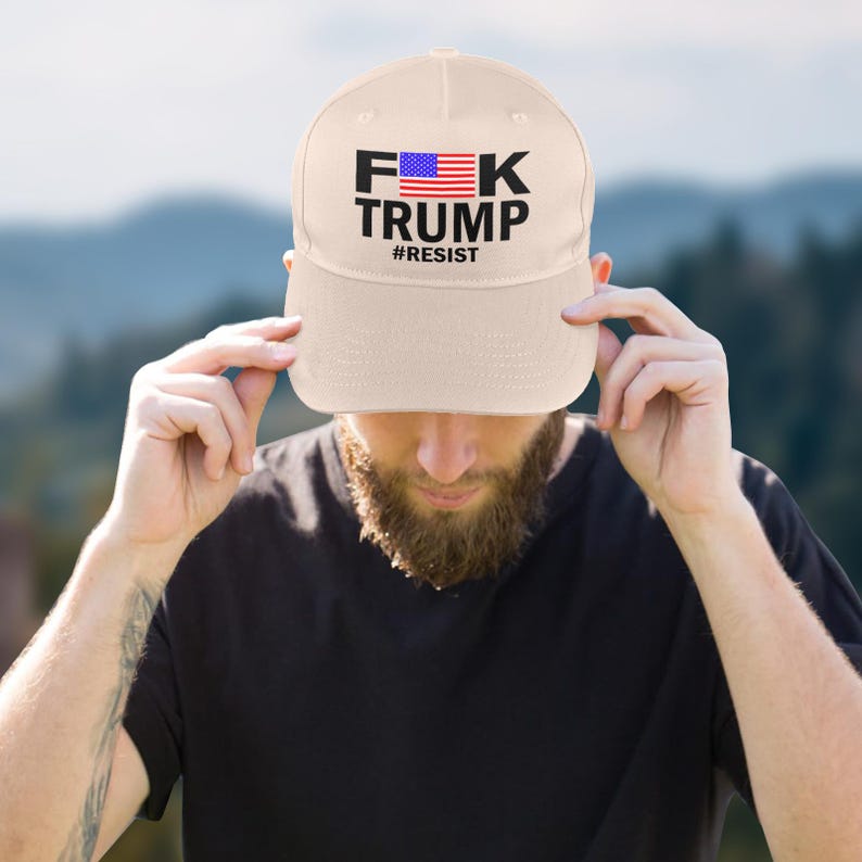 May include: A beige baseball cap with the text "F*K TRUMP" in black letters, with the American flag incorporated into the "F*K" and the word "#RESIST" below. The cap is being held up by a person.