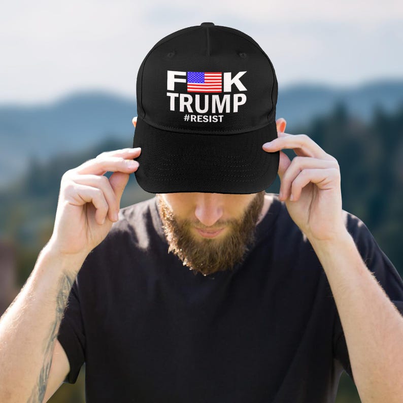 May include: Black baseball cap with the text "F*K TRUMP" in white letters, an American flag graphic, and the word "#RESIST" below. The cap is being held by a person.