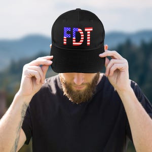 May include: Black baseball cap with the letters "FDT" in a red, white, and blue American flag design. The cap is worn by a person with a beard and a black shirt.