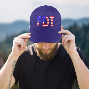 May include: A navy blue baseball cap with the letters "FDT" in white, red, and blue, resembling the American flag. The cap is being held by a person.