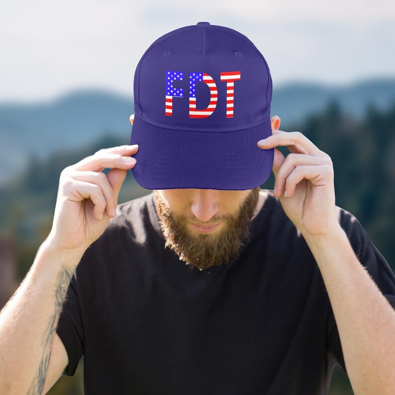 May include: A navy blue baseball cap with the letters "FDT" in white, red, and blue, resembling the American flag. The cap is being held by a person.