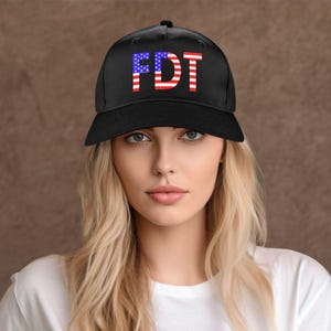 May include: Black baseball cap with the letters "FDT" in red, white, and blue, resembling the American flag. The cap is worn by a person with blonde hair and a white shirt.