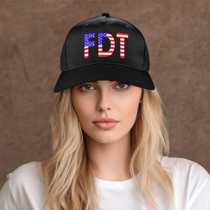May include: Black baseball cap with the letters "FDT" in red, white, and blue, resembling the American flag. The cap is worn by a person with blonde hair and a white shirt.