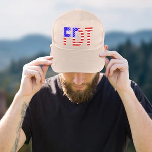 May include: A beige baseball cap with the letters "FDT" in red and blue, designed to resemble the American flag. The cap is being held by a person wearing a black shirt.