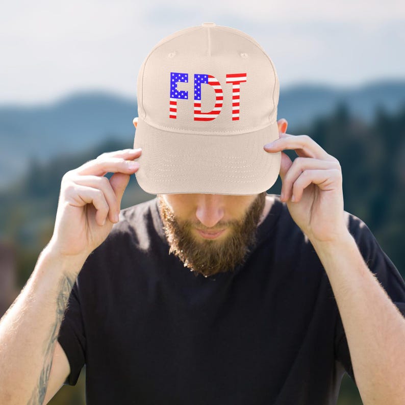 May include: A beige baseball cap with the letters "FDT" in red and blue, designed to resemble the American flag. The cap is being held by a person wearing a black shirt.