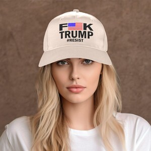 May include: Cream-colored baseball cap with the text "F*K TRUMP" in black block letters, with an American flag graphic and the word "#RESIST" below. The hat is a classic six-panel design.