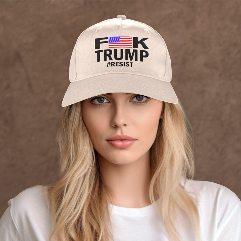May include: Cream-colored baseball cap with the text "F*K TRUMP" in black block letters, with an American flag graphic and the word "#RESIST" below. The hat is a classic six-panel design.