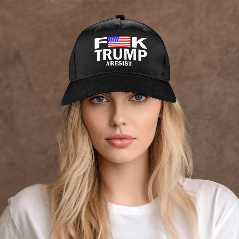 May include: Black baseball cap with the text "F K TRUMP" in white block letters, with an American flag graphic and the word "#RESIST" below. The cap is worn by a person with blonde hair.