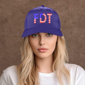 May include: A blue baseball cap with the letters "FDT" in red and white, designed to resemble the American flag. The hat is worn by a person with blonde hair and a white shirt.