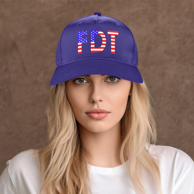 May include: A blue baseball cap with the letters "FDT" in red and white, designed to resemble the American flag. The hat is worn by a person with blonde hair and a white shirt.