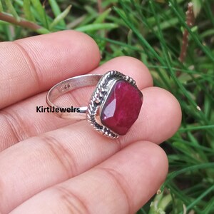 Op de afbeelding: Een zilveren ring met een gefacetteerde, rechthoekige dieprode edelsteen in een decoratieve, gedraaide metalen rand. De binnenkant van de ring is duidelijk gestempeld met het keurmerk 925, wat duidt op een samenstelling van sterling zilver.