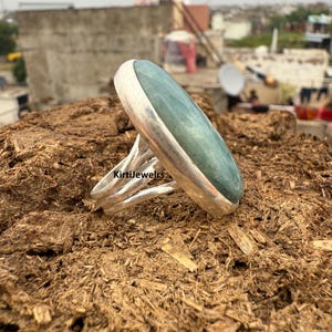 Può includere: Un anello d'argento con una grande pietra ovale verde pallido. La fascia dell'anello ha un design strutturato e aperto. L'anello è esposto su una superficie marrone e strutturata. Il testo "KirtiJewelrs" è visibile sull'anello.