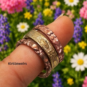 May include: A set of three stacked rings featuring two copper-toned bands with embossed floral and heart patterns and one central gold-toned band with an engraved sunburst design. The text KirtiJewelrs is visible on the side of the rings.