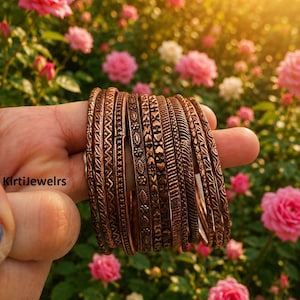 May include: A set of ten copper-toned metal bangles featuring various embossed geometric, floral, and heart patterns. The bracelets have a dark, oxidized finish and are stacked together. The text KirtiJewelrs is visible on the left side.