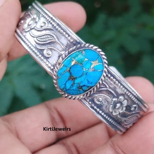 May include: A silver cuff bracelet featuring an oval turquoise stone with dark matrix patterns in the center, surrounded by a twisted rope border. The band is decorated with embossed floral and leaf motifs and the text KirtiJewelrs.