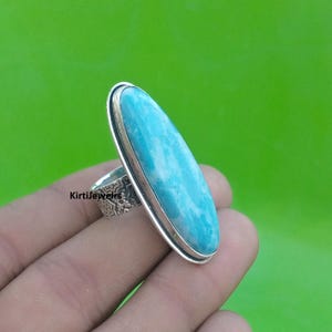May include: A silver ring featuring a large, oval-shaped, light blue gemstone with white marbling. The gemstone is set in a silver bezel. The ring band has an ornate, textured design. The background is a solid green.
