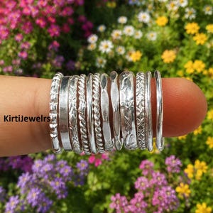 May include: A collection of eleven silver rings with various textures and patterns, including twisted, smooth, and engraved designs, arranged side by side. The text KirtiJewelrs is visible on the left side of the silver ring collection.