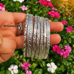 May include: A set of six silver-toned metal bangles featuring various embossed patterns including floral motifs, wavy lines, and geometric shapes, with the text KirtiJewelrs visible on the side of the bangles.