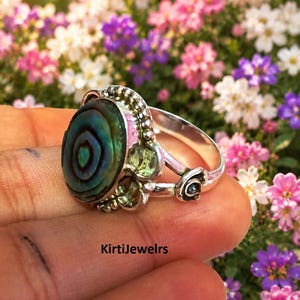Può includere: Un anello in argento con un grande cabochon ovale in conchiglia di abalone dai toni iridescenti blu e verdi. La montatura include piccole pietre preziose sfaccettate verde chiaro e decorazioni in metallo a perline. Il testo riporta KirtiJewelrs.