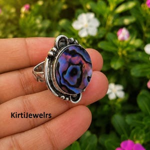 Könnte beinhalten: Ein Silberring mit einer großen, ovalen Abalone-Muschel-Einlage mit irisierenden violetten, blauen und schwarzen Wirbelmustern. Die Metallfassung hat ein dekoratives, oxidiertes Finish mit kleinen Perlenakzenten. Der Text KirtiJewelrs ist sichtbar.