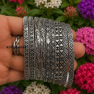 Op de afbeelding: Een set van acht zilverkleurige metalen armbanden met diverse reliëfpatronen, waaronder bloemranken, geometrische ruiten en hartvormen. De armbanden hebben een geoxideerde afwerking en de tekst KirtiJewels is op de afbeelding zichtbaar.