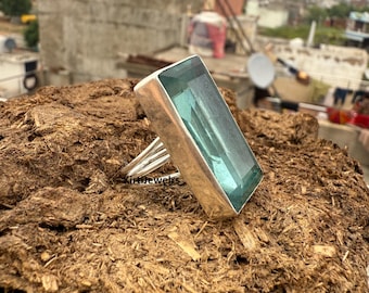 Puede incluir: Anillo rectangular de plata con una piedra preciosa azul claro. La piedra está engastada en un bisel de plata y tiene un aspecto claro y transparente. El anillo se muestra sobre una superficie marrón texturizada.