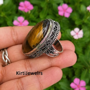 Puede incluir: Un anillo con una piedra preciosa ojo de tigre ovalada engastada en una banda de plata decorativa con intrincados trabajos de filigrana. La piedra presenta bandas chatoyantes marrones y doradas. El texto KirtiJewelrs es visible.