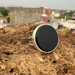 May include: A silver ring with a large, round, black onyx stone. The ring has a thick band and a textured design on the sides. The stone is set in a bezel setting. The ring is displayed on a brown, textured surface.
