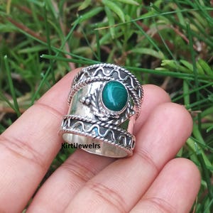 May include: A wide silver ring featuring a central oval green malachite gemstone. The band is decorated with two horizontal rows of twisted rope patterns and wavy lines. The text KirtiJewelrs is printed in black on the lower part of the band.