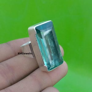 May include: A rectangular, teal gemstone ring with a silver band. The gemstone is set in a silver bezel and has a clear, glassy appearance. The ring is displayed against a green background. The text "KirtiJewelrs" is visible on the ring.